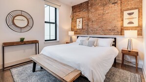 A bedroom with a large bed, a wooden bench, a round wall decoration, a brick wall, and a window. - Modern Condo in the heart of Over-The-Rhine (Cincinnati)
