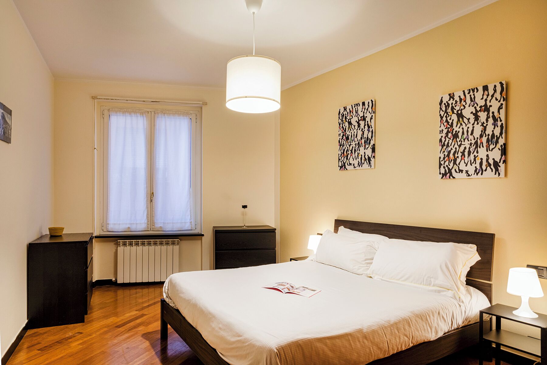 Une chambre d’hôtel comprenant un lit, des tables de chevet, une fenêtre avec des rideaux, un radiateur et deux œuvres d’art murales.