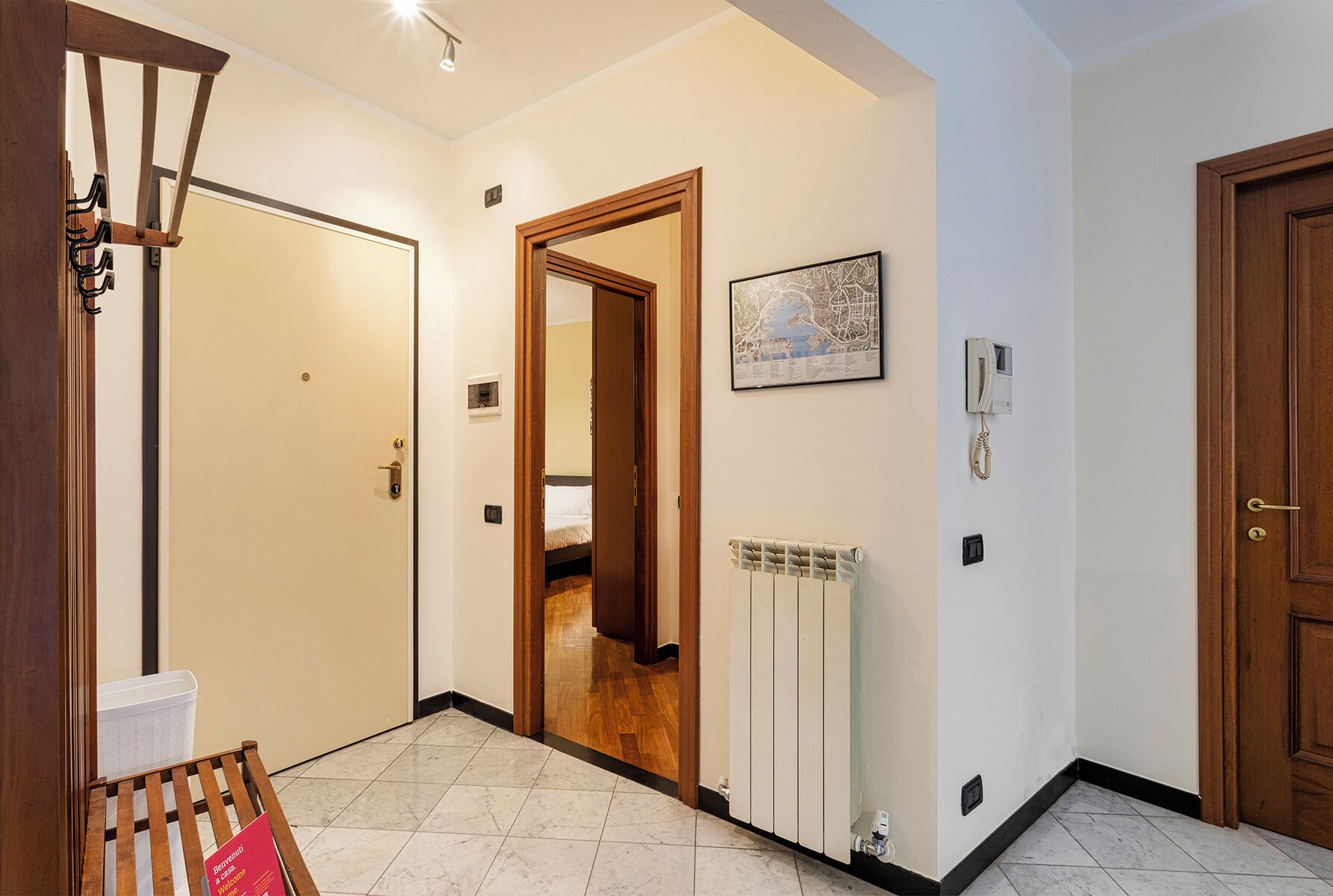 Un couloir d’hôtel avec un banc en bois, un radiateur, une carte encadrée et une porte donnant sur une chambre.