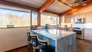 Une cuisine moderne dotée d’un grand îlot central, de tabourets de bar et offrant une vue sur les arbres grâce à de grandes fenêtres.