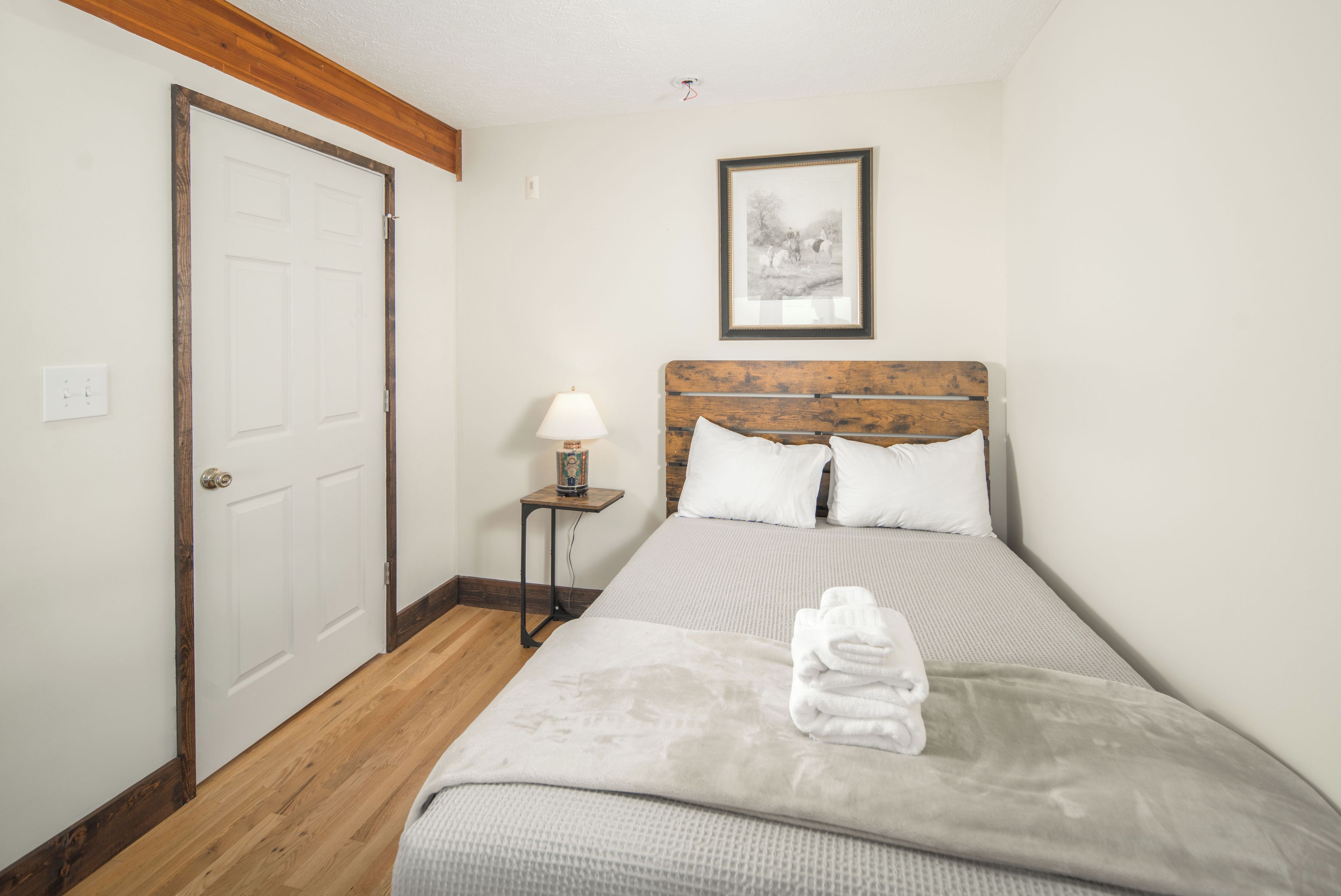 A bedroom with a wooden headboard, a bed with white bedding, a bedside table with a lamp, and a framed picture on the wall.