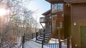 Une cabane en bois à deux étages, dotée d’un balcon, d’un escalier et d’une grande fenêtre donnant sur une forêt enneigée.