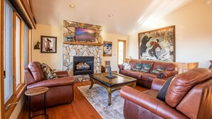 A cozy living room with leather sofas, a stone fireplace, and artwork on the walls. - 5,000ft Peak View Home: Hot Tub, Fire Table & 4 Kings; 5 Min to Resort (Beech Mountain)