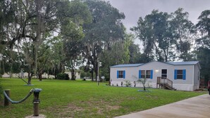 A small, single-story house with a blue door and windows, surrounded by a grassy yard and trees. - Cozy 3-bedroom, 2 full bath mobile home on half acre in charming Welaka! (Welaka)