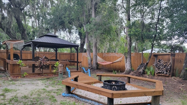A backyard with a wooden deck, a hammock, a fire pit, and a covered seating area. - Cozy 3-bedroom, 2 full bath mobile home on half acre in charming Welaka! (Welaka)