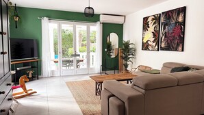 A living room with a sofa, a TV, a dining table, and a large window. - Villa Zanoli - a tropical paradise with swimming pool, close to everything! (Appartement)