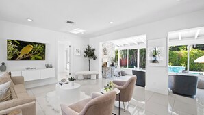 Una sala moderna con una decoración blanca, un televisor de pantalla plana grande, un banco y vistas al exterior a través de ventanas amplias.