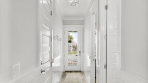 A narrow hallway with white walls and doors, leading to a glass door with a view of the outside.