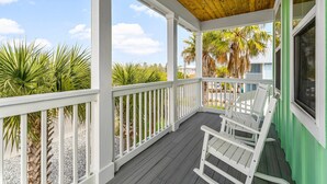 A covered porch with white railings, a ceiling fan, and a view of palm trees and a building in the background.