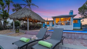 A poolside area with lounge chairs and a thatched umbrella, a pool, and a building with a blue exterior and a visible staircase. - Gulf Access, Heated Pool & Spa, Tiki Hut - Roelens (Fort Myers Beach)