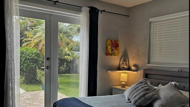 Ein Schlafzimmer mit einem Bett, einem Deckenventilator, einem Fenster mit Jalousien und Blick auf Grünfläche.
