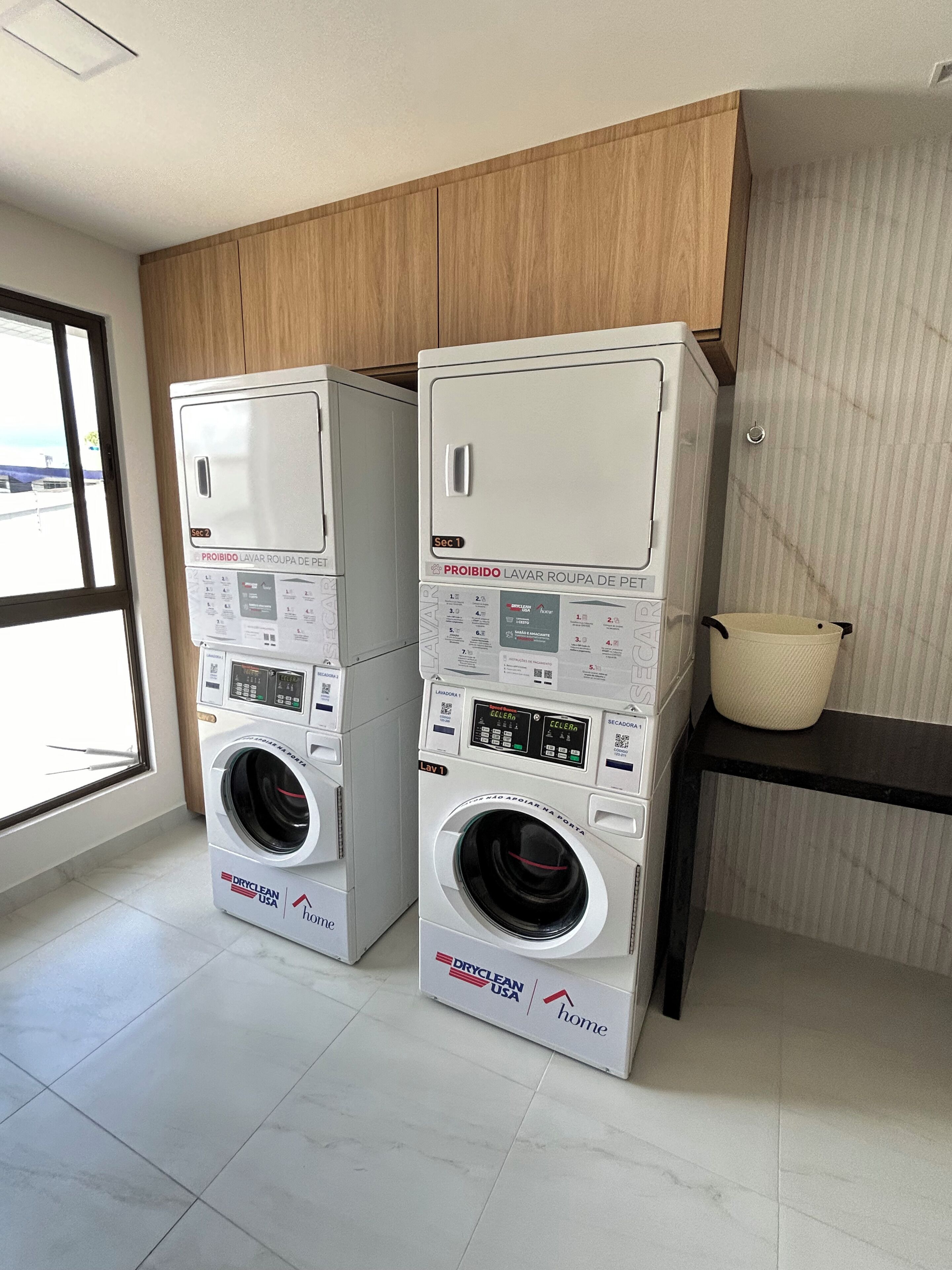 Two stacked front-loading washing machines in a laundry room with a window and a wooden cabinet above.