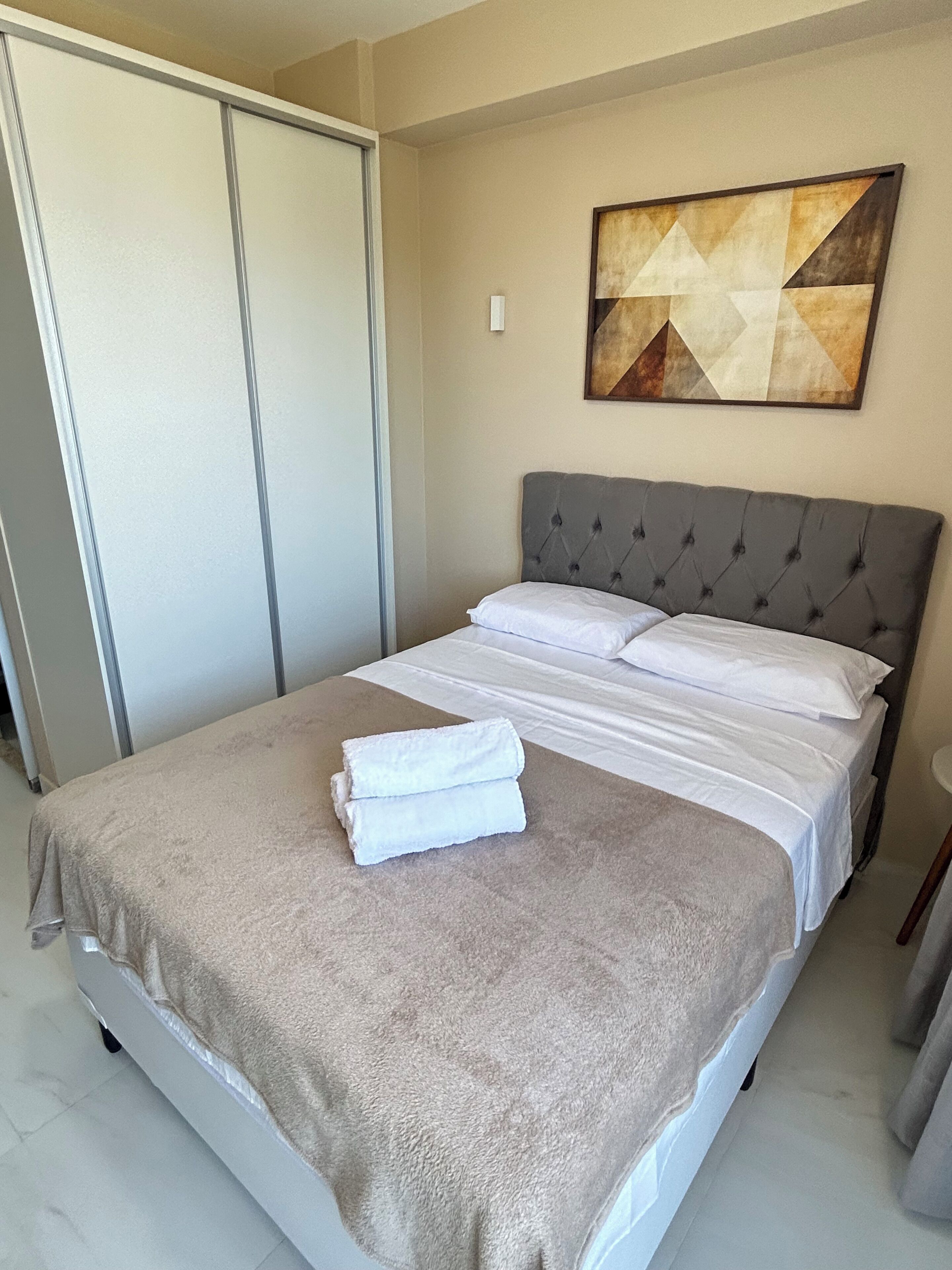 A neatly made bed with a grey headboard and white linens, a beige blanket, and two folded white towels on the bed.