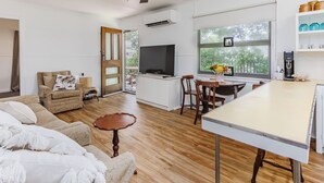 A modern living space with a dining area, kitchen, and a large window offering a view of greenery. - Cozy Pet Friendly Beach House (Kingscliff)