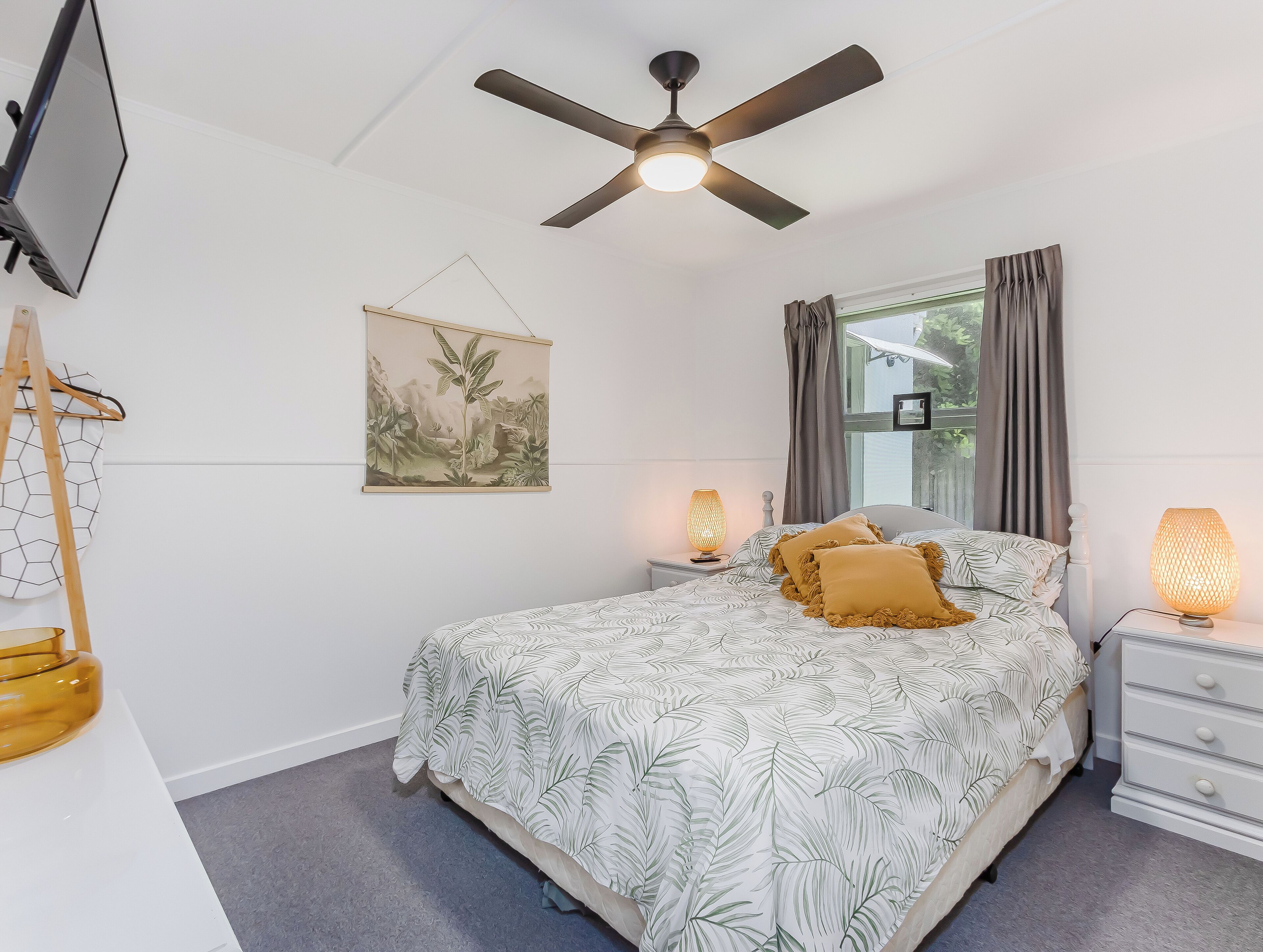 Un dormitorio con cama, ventilador de techo, televisor montado en la pared, ventana con cortinas y lámpara de noche.