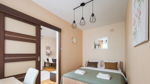 A bedroom with a bed, bedside table, and a wooden wardrobe. There is a dining area visible through an open door.