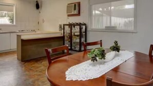 Un comedor con una mesa de madera, sillas y un mantel de encaje. Se observa un ventilador de techo, una ventana y una zona de cocina al fondo.