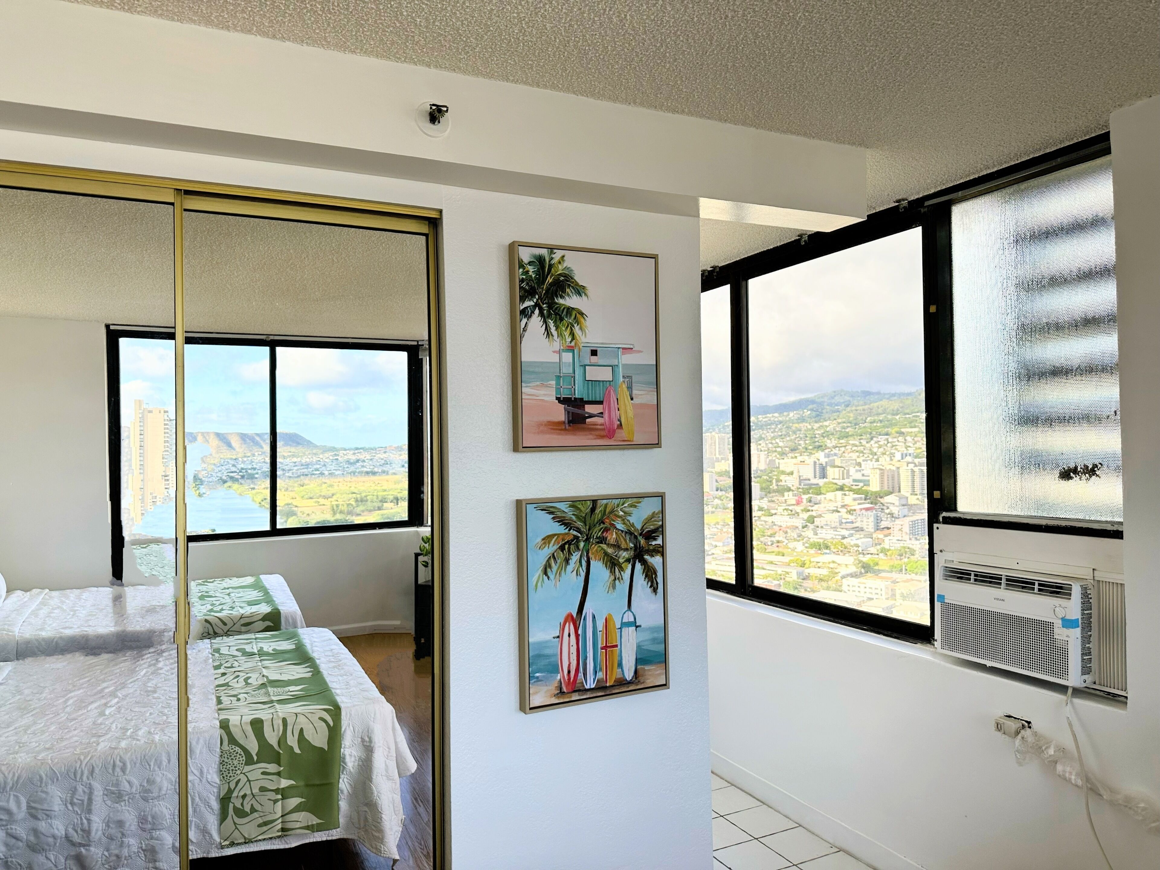 A bedroom with a bed, a large window with a city view, and two wall-mounted pictures.