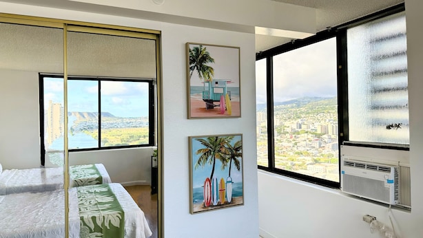 A bedroom with a bed, a large window with a city view, and two wall-mounted pictures. - Waikiki Studio with Stunning Views! (Honolulu)