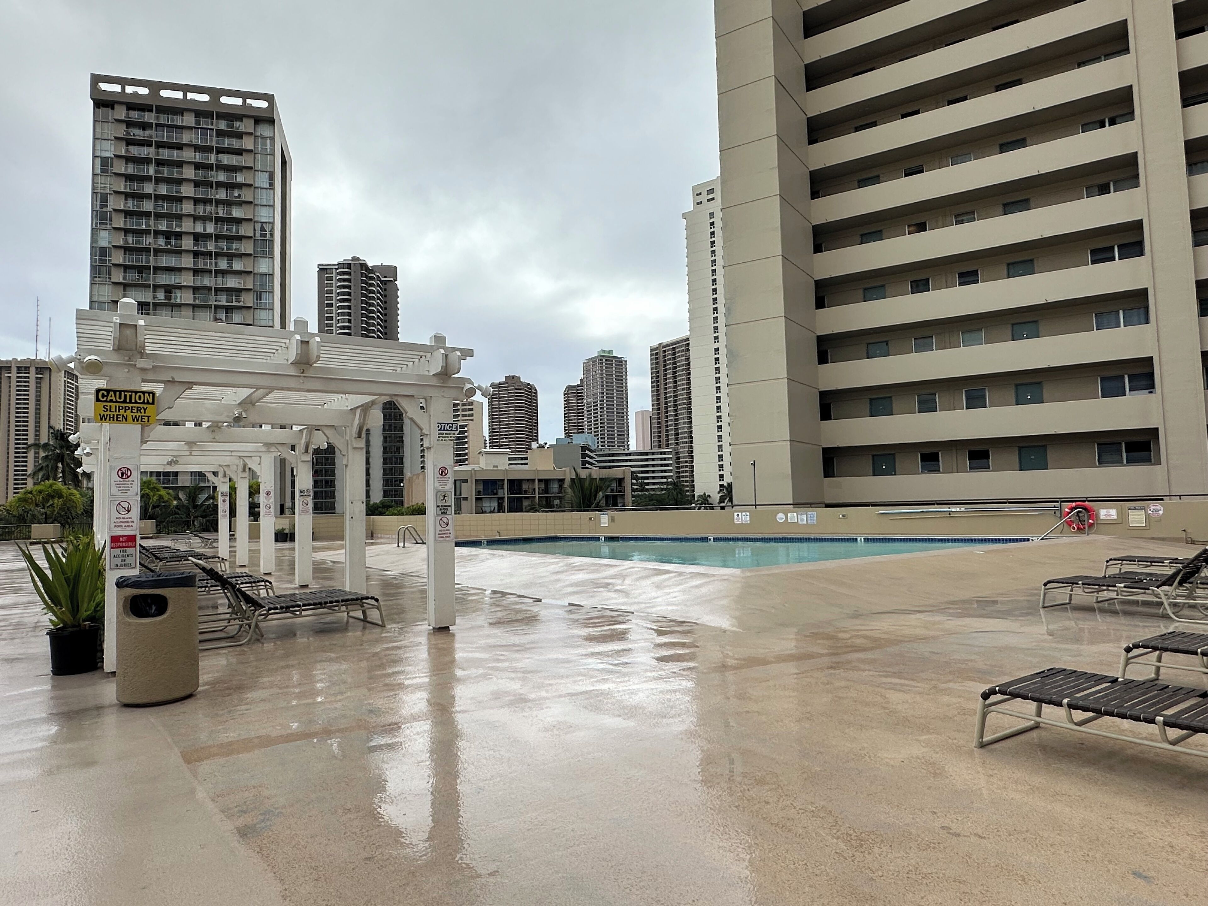 A rooftop pool area with a white pergola, lounge chairs, and a caution sign for slippery surfaces when wet.