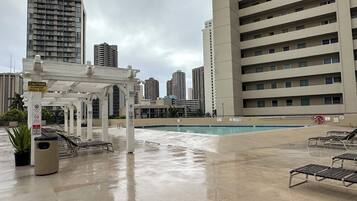 A rooftop pool area with a white pergola, lounge chairs, and a caution sign for slippery surfaces when wet.