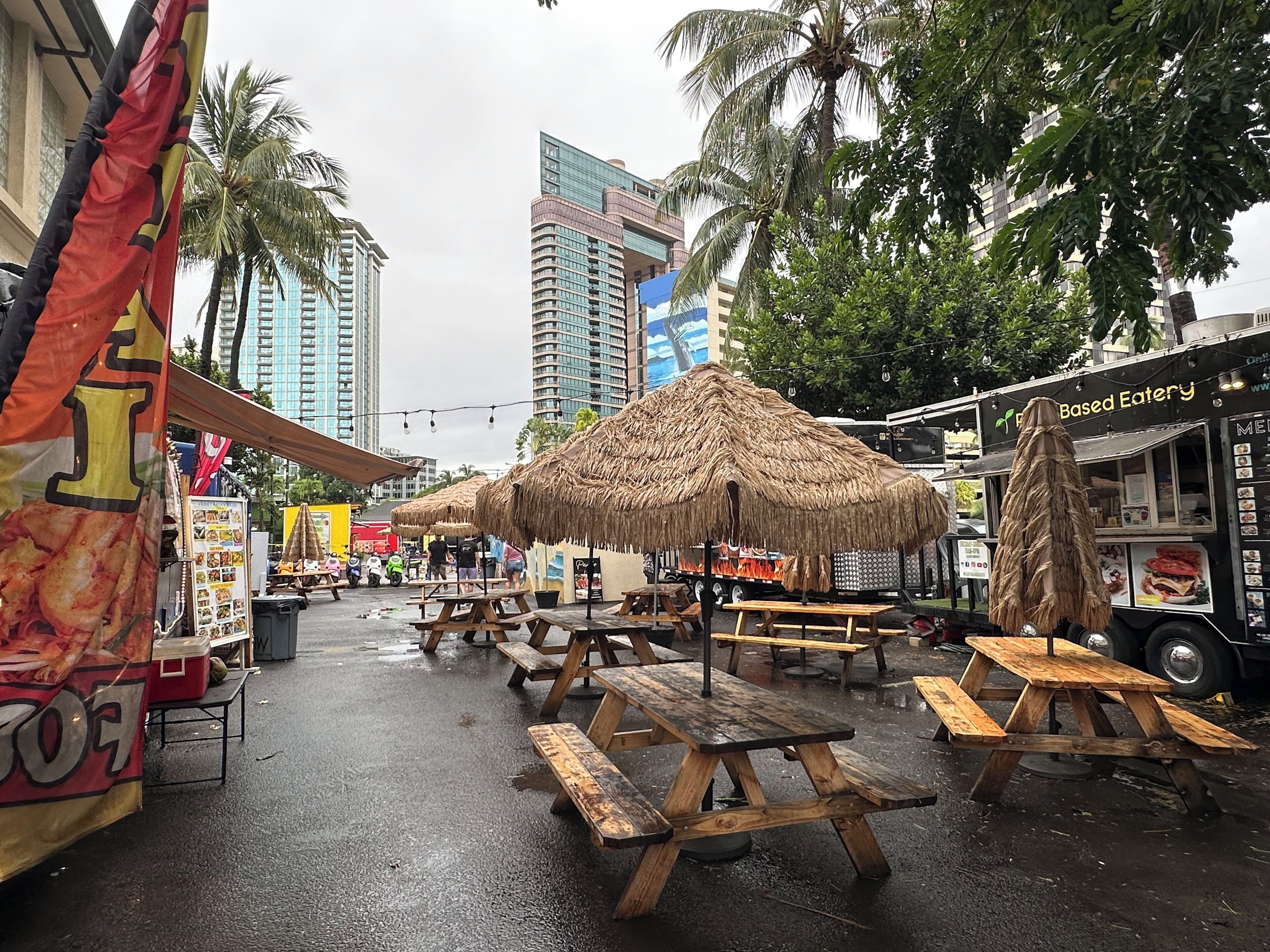 An outdoor dining area with wooden picnic tables and thatched umbrellas, surrounded by tall buildings and palm trees.