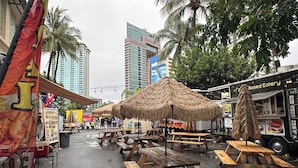 An outdoor dining area with wooden picnic tables and thatched umbrellas, surrounded by tall buildings and palm trees. - Waikiki Studio with Stunning Views! (Honolulu)