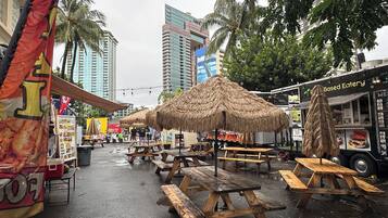 An outdoor dining area with wooden picnic tables and thatched umbrellas, surrounded by tall buildings and palm trees.