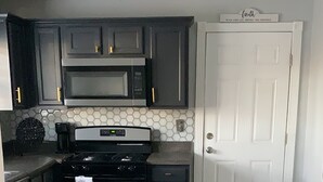 Una cocina con armarios oscuros, un microondas, una estufa y un lavavajillas. Hay una puerta blanca y un candelabro en el techo.