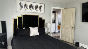 Un dormitorio con cama negra, cabecera con marco dorado, puerta blanca y televisión montada en la pared.