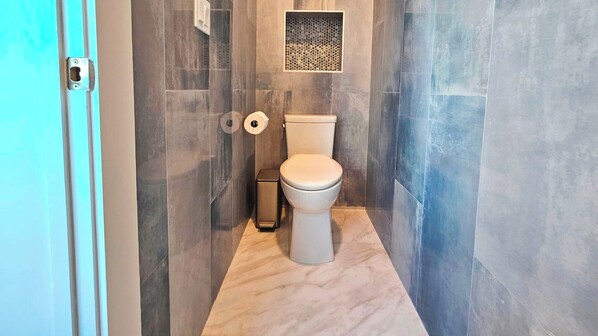 A bathroom with a toilet, a trash bin, and a shower area.