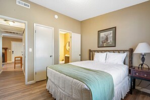 A bedroom with a bed, a bedside table, a lamp, a framed picture on the wall, and an open door leading to another room.