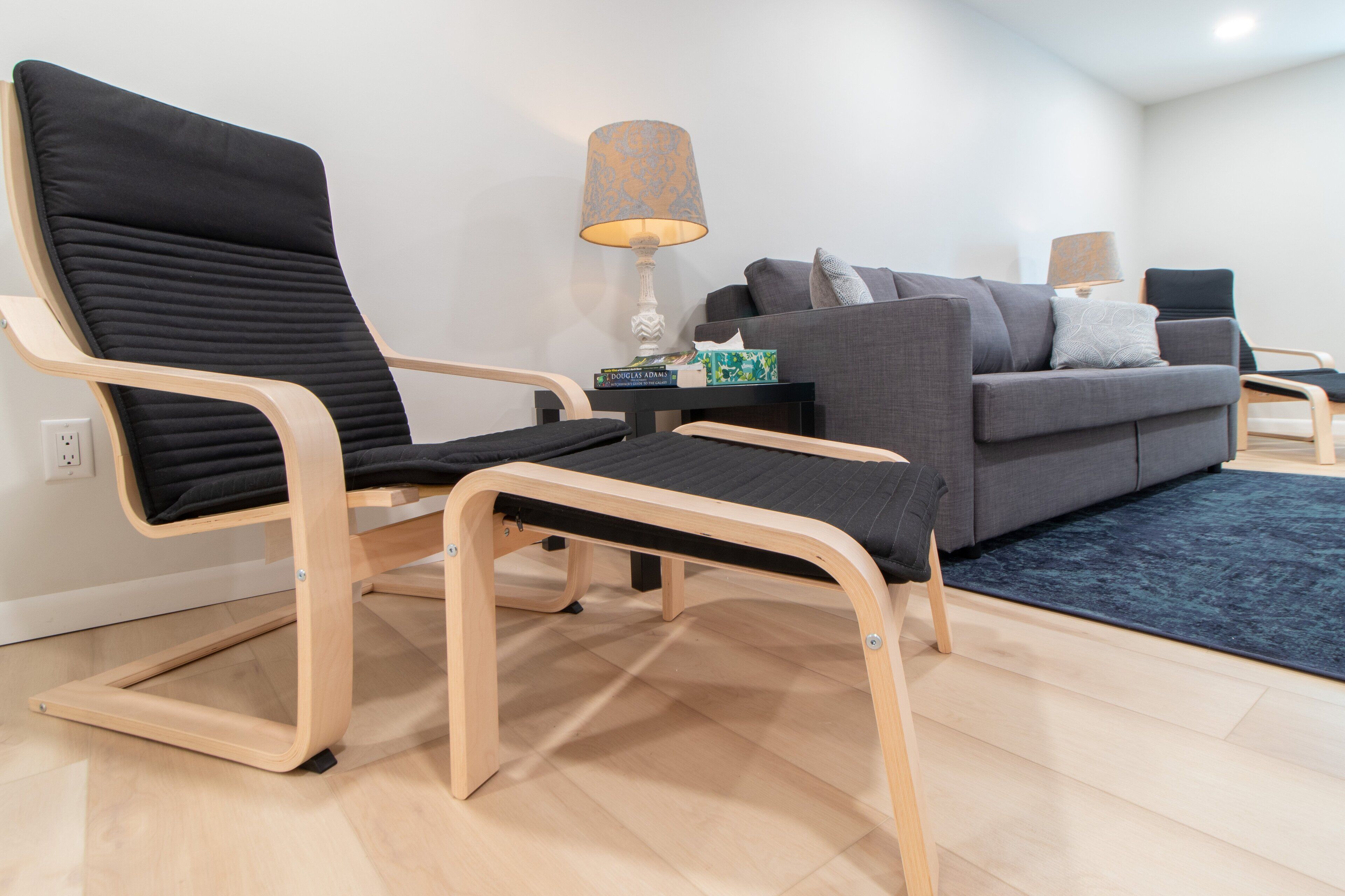 A modern living room with a wooden-framed sofa, a black cushioned chair, and a small side table with a lamp and books.