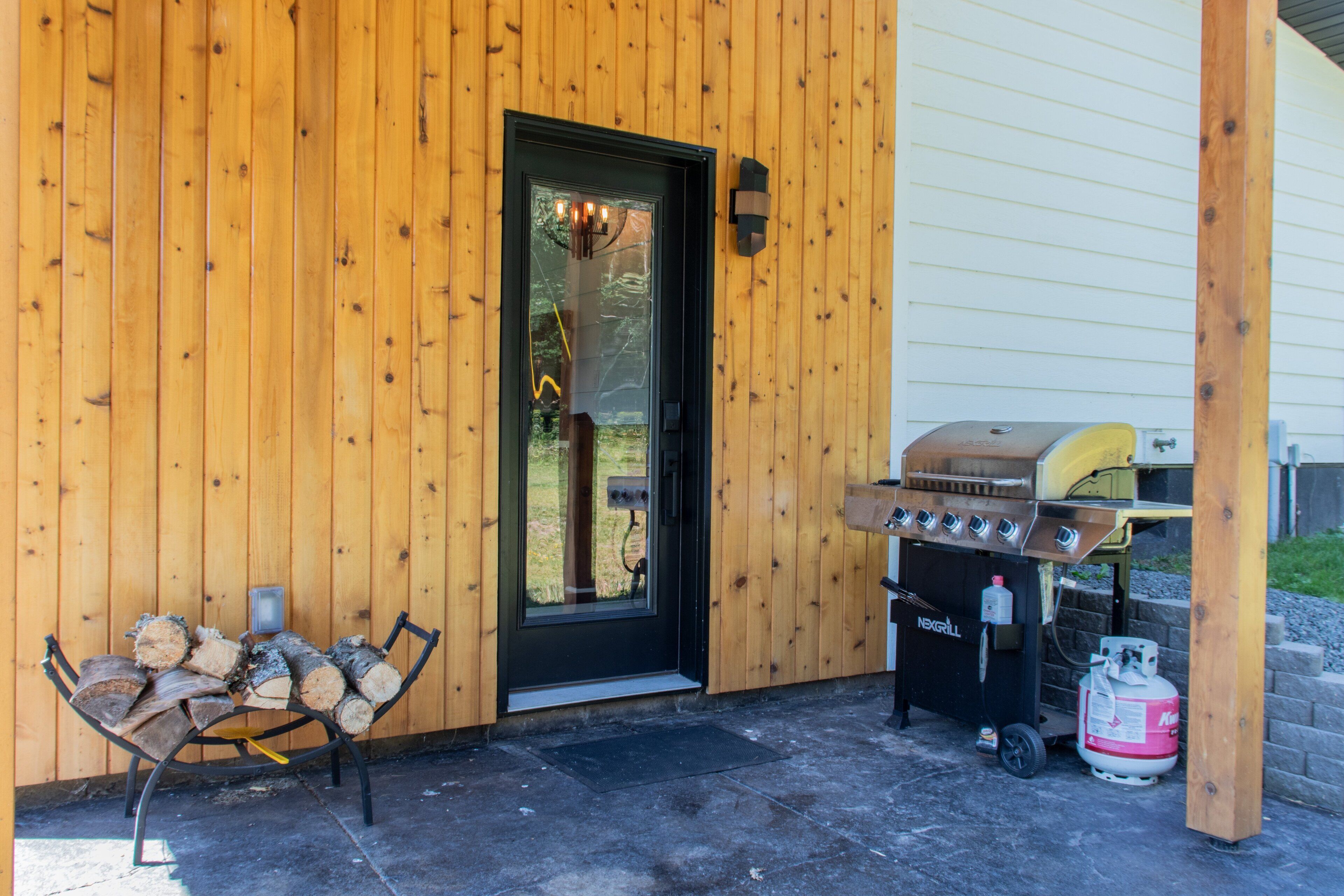 Fachada de madera con una puerta negra, una parrilla y un banco con leña.