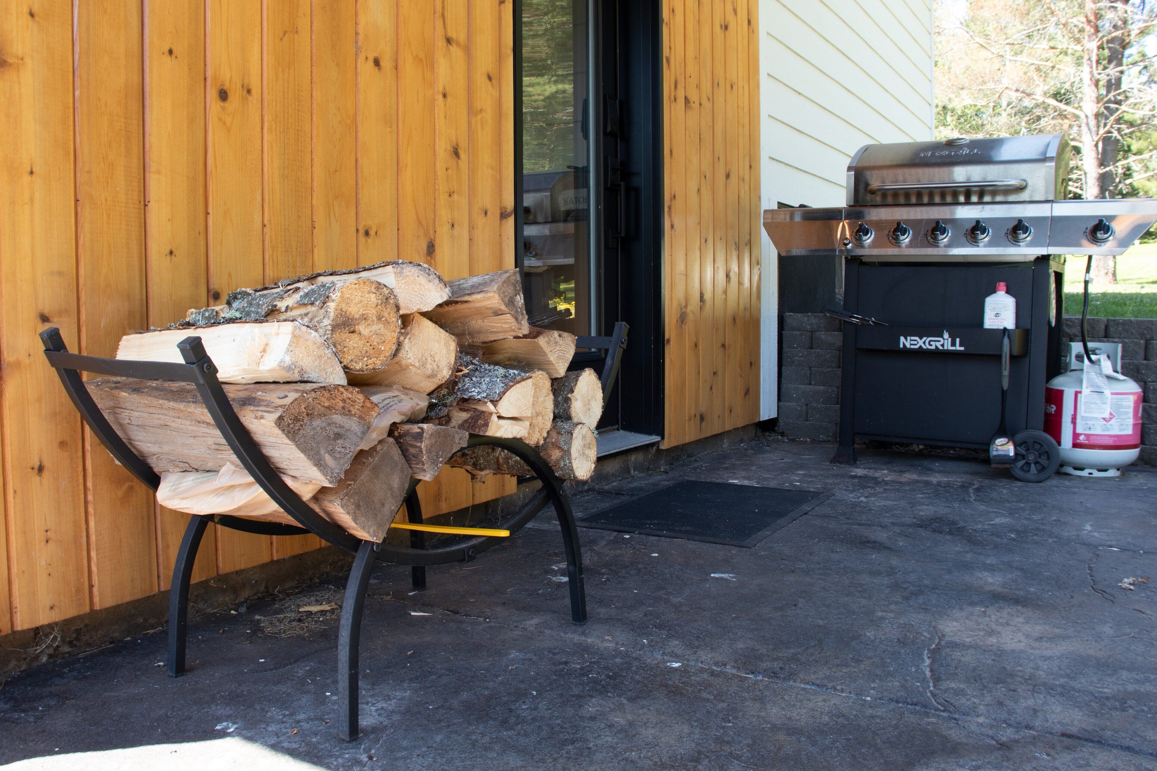 Pared exterior de madera, una pila de leña y una parrilla portátil con tanque de gas.