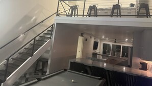 A modern interior with a pool table, a staircase, and a kitchen area.