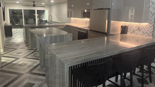 A modern kitchen with a marble island, black bar stools, and a herringbone-patterned floor.