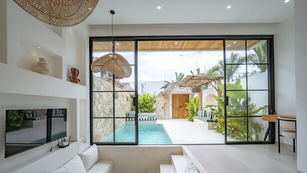 A modern living room with a large glass door leading to a pool area, white sofas, a flat-screen TV, and a woven ceiling light. - Villa Ratu Canggu (Canggu)