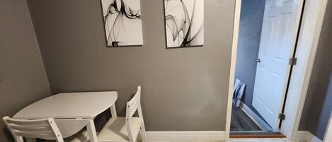 A room with a white table and chairs, two abstract paintings on the wall, and a door leading to a hallway.