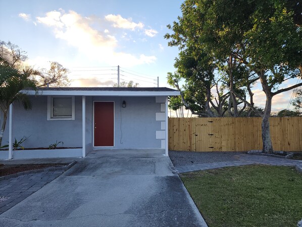 Una casa pequeña de un piso con puerta roja, un ventanal y una valla de madera.