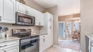 A kitchen with white cabinets, a stainless steel microwave, and a stove. There is a dining area with a table and chairs visible through an open door. - 7028 Ocean Lakes Golf Cart⛱️🏝️🩱🌊👙☀️🐚 (Myrtle Beach)