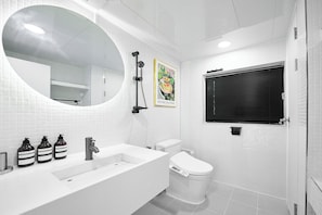 Une salle de bain moderne avec un lavabo, des toilettes et une douche, le tout de couleur blanche. On peut voir trois flacons de savon liquide sur le lavabo.