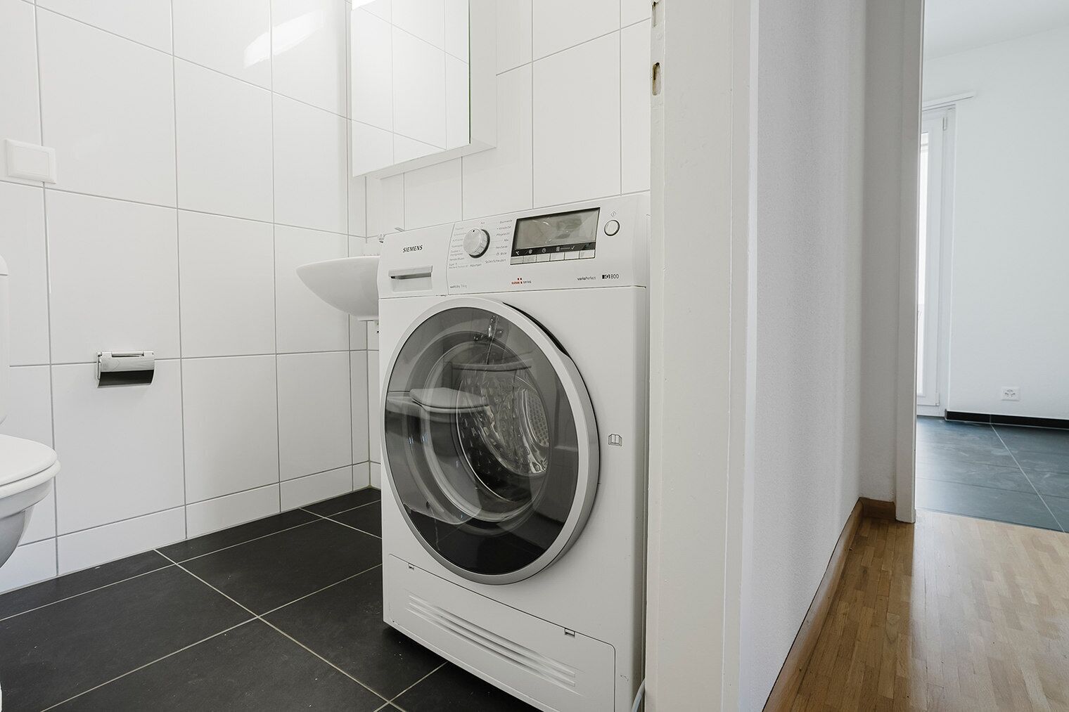 Et moderne badeværelse med en hvid flisevæg, en sort flisegulv og en Siemens vaskemaskine.