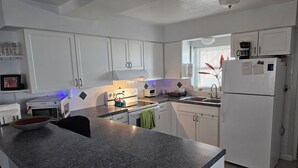 A modern kitchen with white cabinets, a granite countertop, a refrigerator, and a window with blinds.