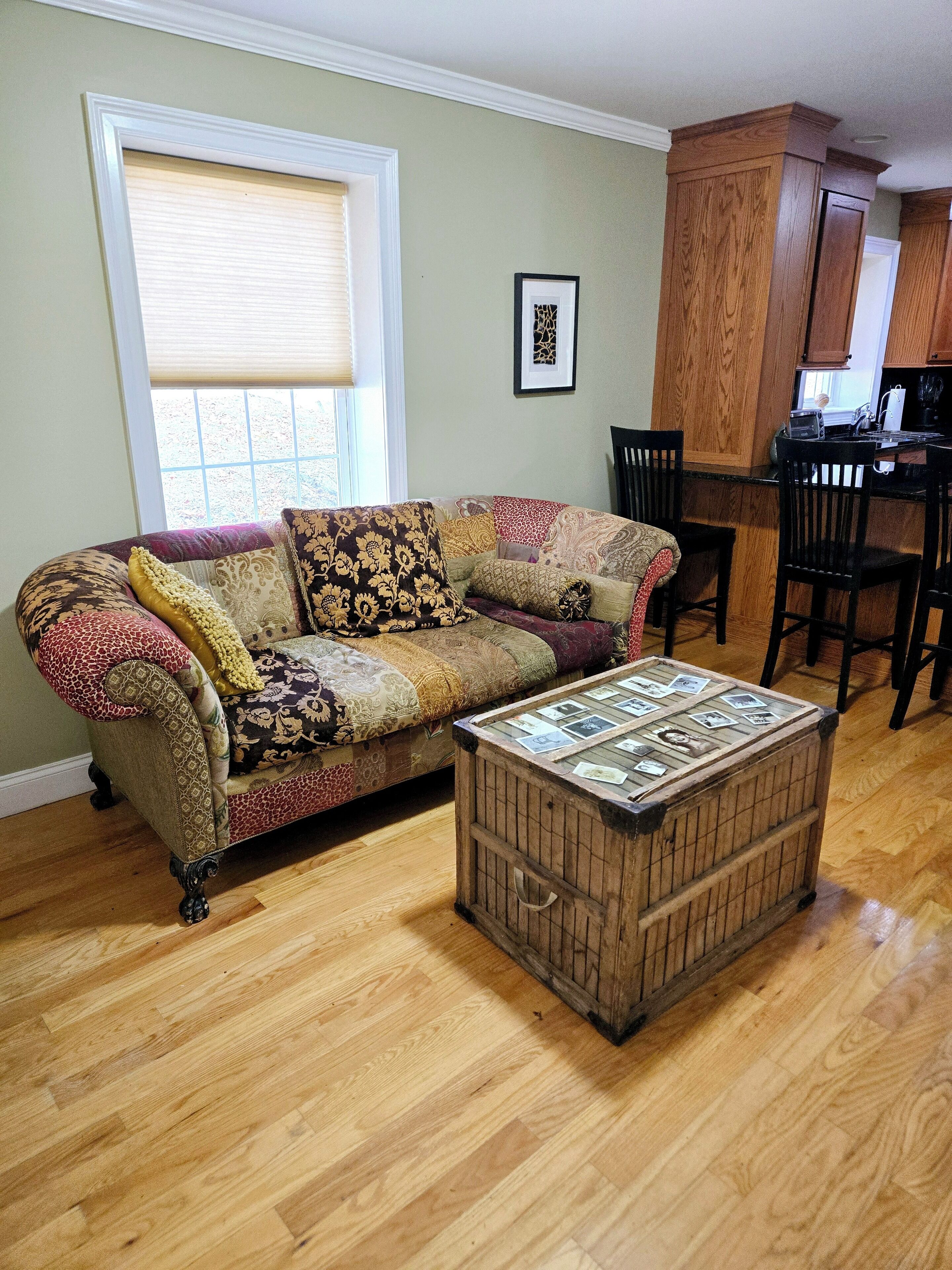 A living room with a sofa, a coffee table with photos, wooden flooring, and a kitchen area with wooden cabinets and chairs.