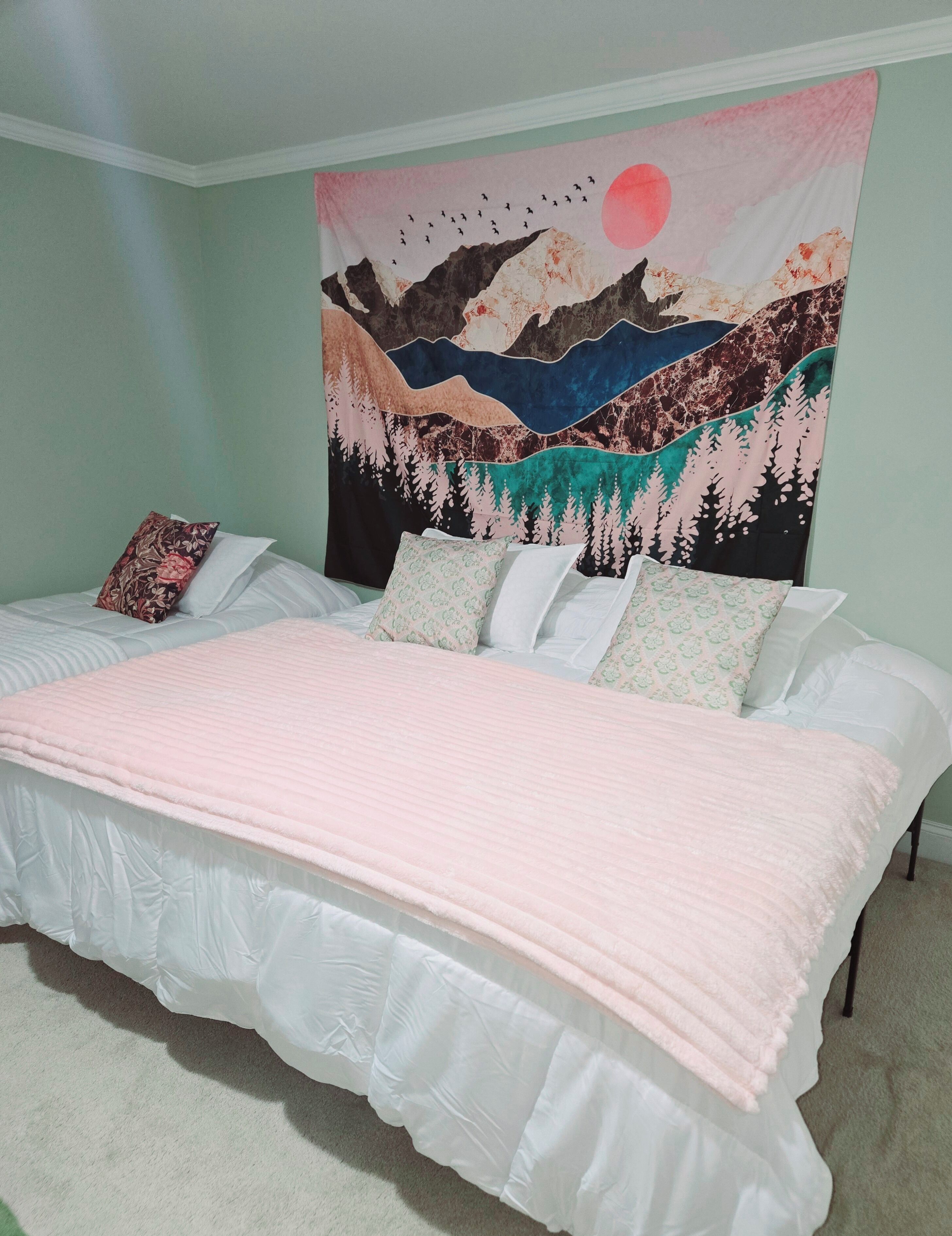 A bedroom with a bed, pillows, and a tapestry depicting a landscape with mountains and a sun.
