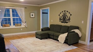 Ein Wohnzimmer mit Ecksofa, einem weißen Teppich, Holzböden, einem Fenster mit Vorhängen und einer Wanddekoration.