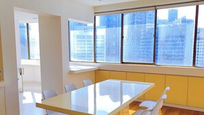 Ein moderner Essbereich mit einem großen rechteckigen Tisch und sechs weißen Stühlen. Der Raum verfügt über Holzböden, große Fenster mit Blick auf die Stadt und eine helle, freundliche Atmosphäre.