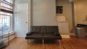 Ein Wohnzimmer mit einem dunkelbraunen Sofa, einer weißen Tür, einem Fenster mit Jalousien, einem Heizkörper und einer kleinen Mülltonne.