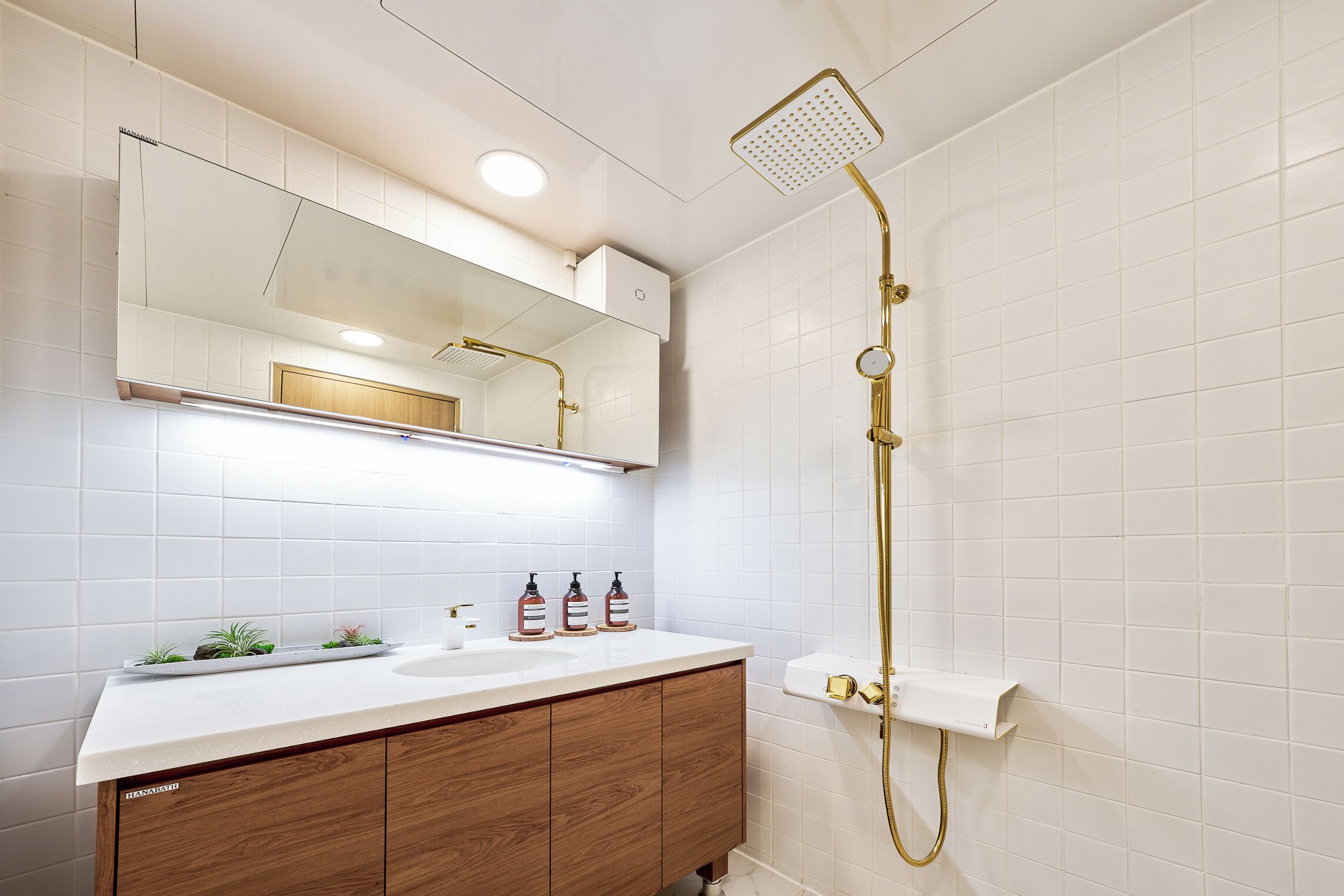 Une salle de bain avec un mur recouvert de carreaux blancs, un meuble-lavabo en bois, un grand miroir et une pomme de douche dorée.